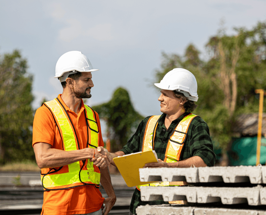 Dois profissionais da construção cumprimentam-se num estaleiro de obras, com capacetes de proteção e equipamento de segurança.