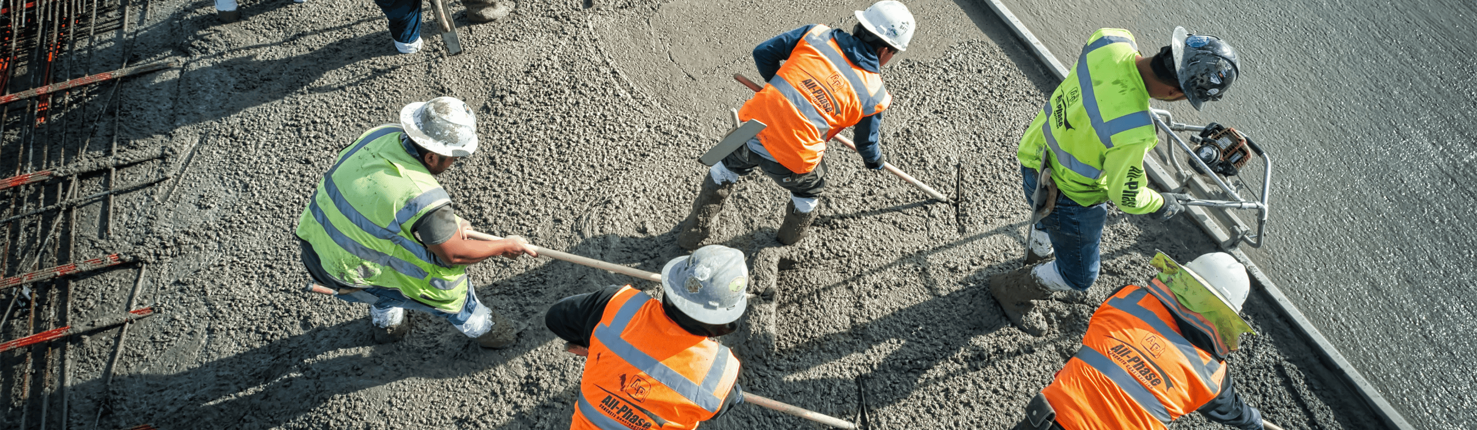 Equipa de trabalhadores da construção com coletes de alta visibilidade e capacetes a trabalhar betão fresco numa grande obra.